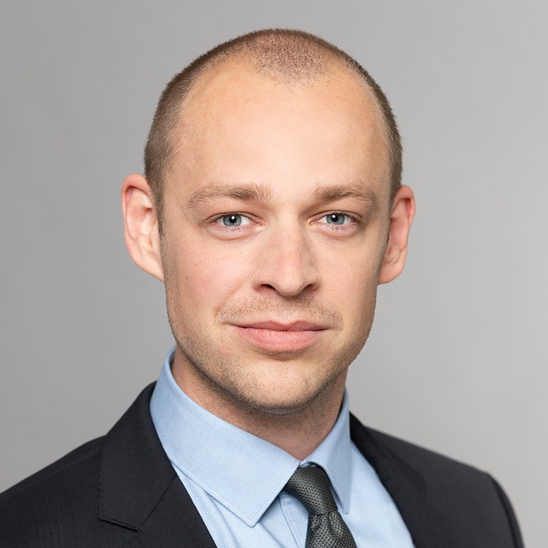 Porträt eines Mannes mit kurzen, hellen Haaren. Er trägt einen dunklen Anzug, ein hellblaues Hemd und eine graue Krawatte. Er lächelt leicht in die Kamera, mit einem neutralen Hintergrund.