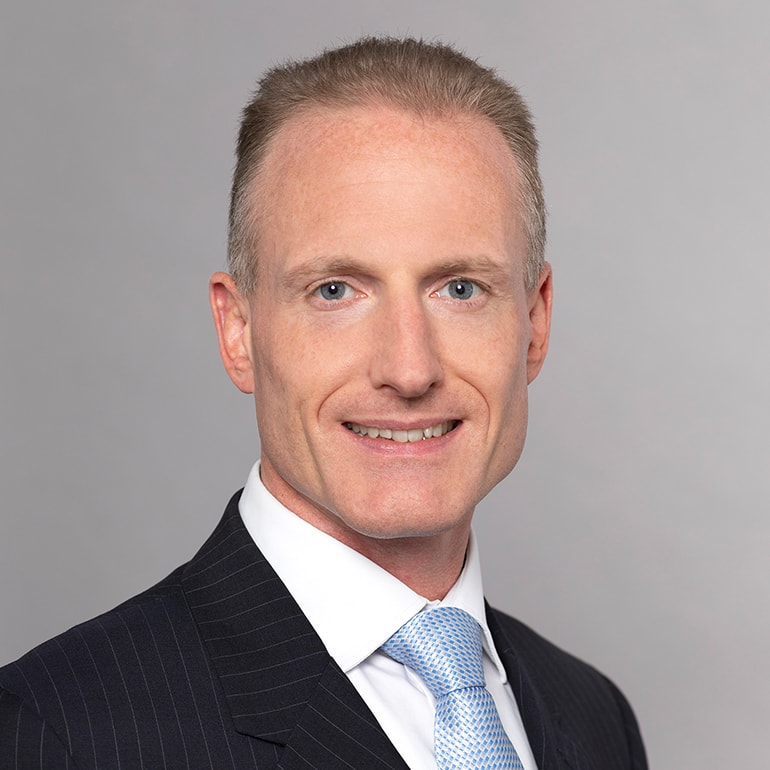 Ein Mann mit kurzen, blonden Haaren und einem freundlichen Lächeln trägt einen dunkel gestreiften Anzug und ein hellblaues Hemd mit einer passenden Krawatte. Er steht vor einem neutralen grauen Hintergrund.