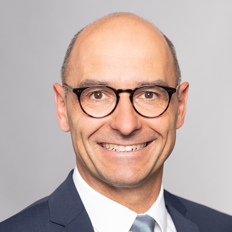 Ein lächelnder Mann mit Glatze trägt eine Brille und einen Anzug. Der Hintergrund ist neutral grau. Der Mann strahlt Selbstbewusstsein und Freundlichkeit aus.