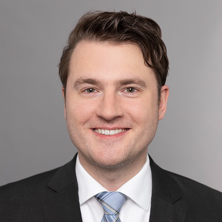 Ein junger Mann mit einem freundlichen Lächeln trägt einen Anzug und eine gestreifte Krawatte. Der Hintergrund ist grau.