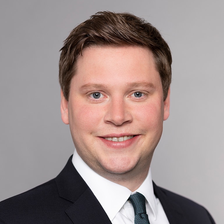 Porträt eines lächelnden Mannes mit kurzen, braunen Haaren. Er trägt einen schwarzen Anzug mit weißem Hemd und einem dunkelgrünen Krawatte. Der Hintergrund ist einfarbig grau, was den Fokus auf sein freundliches Gesicht lenkt.