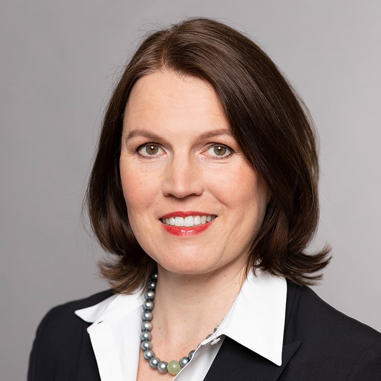 Porträt einer Frau mit mittellangen, braunen Haaren. Sie trägt eine schwarze Jacke und eine weiße Bluse sowie eine Perlenkette. Die Frau lächelt freundlich in die Kamera, mit einem neutralen Hintergrund.