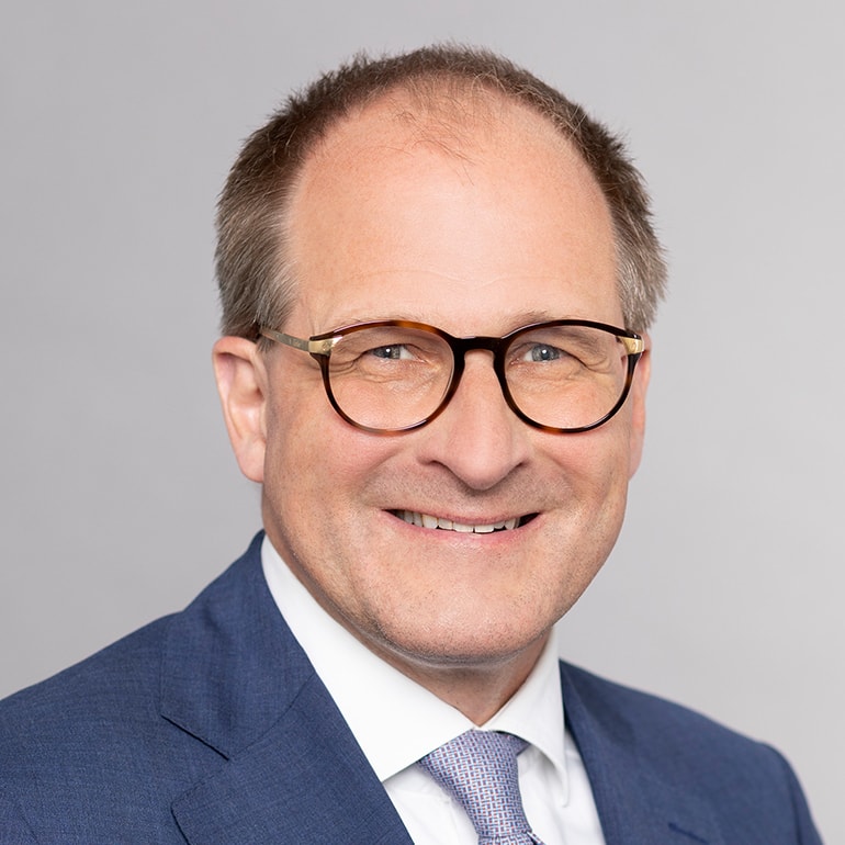 Ein lächelnder Mann mit Brille und einem Anzug in Blau. Der Hintergrund ist hellgrau. Die Halspartie und das Gesicht sind gut sichtbar, was dem Bild einen professionellen Eindruck verleiht.