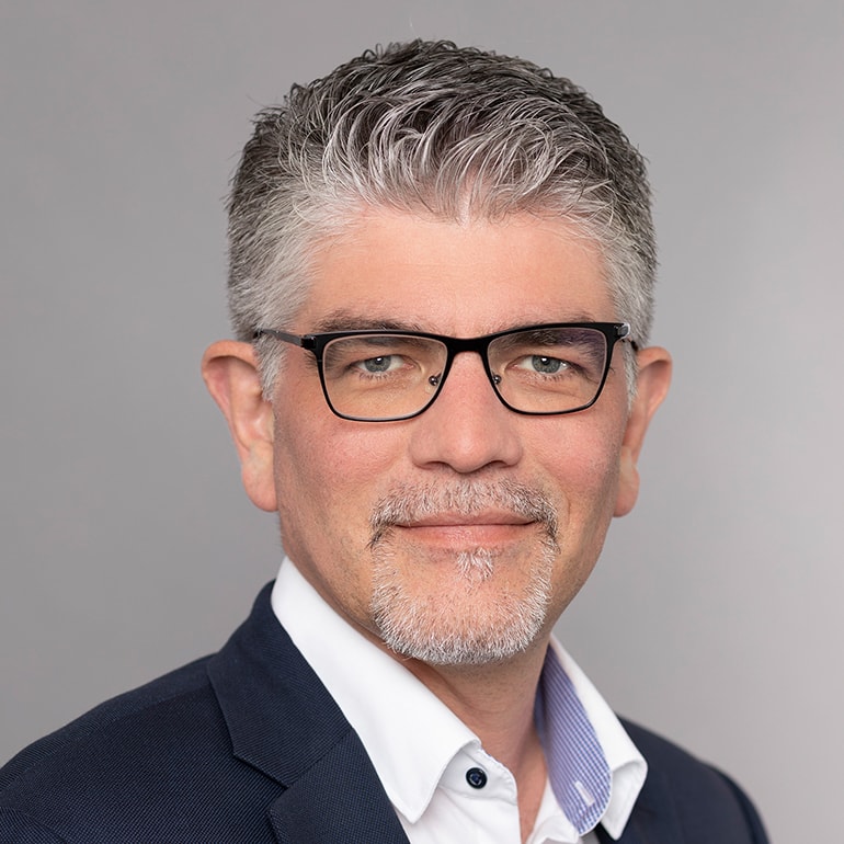 Porträt eines Mannes mittleren Alters mit grauem Haar und Bart. Er trägt eine Brille und einen schwarzen Anzug über einem weißen Hemd mit blauer Krawatte. Der Hintergrund ist neutral grau. Der Mann lächelt freundlich in die Kamera.