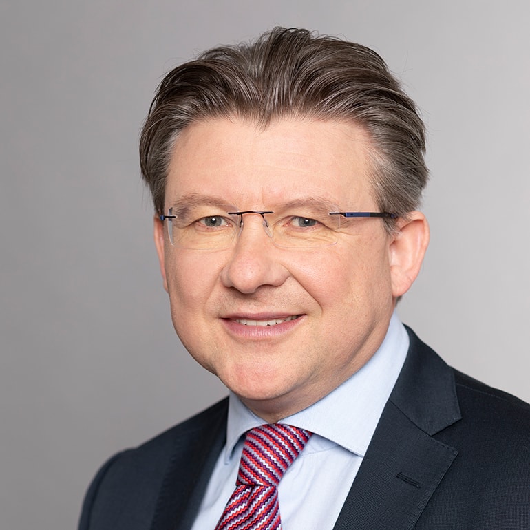 Ein Mann mit braunen, kurzen Haaren und einer Brille lächelt in die Kamera. Er trägt einen Anzug mit einem hellblauen Hemd und einer gestreiften Krawatte. Der Hintergrund ist grau.