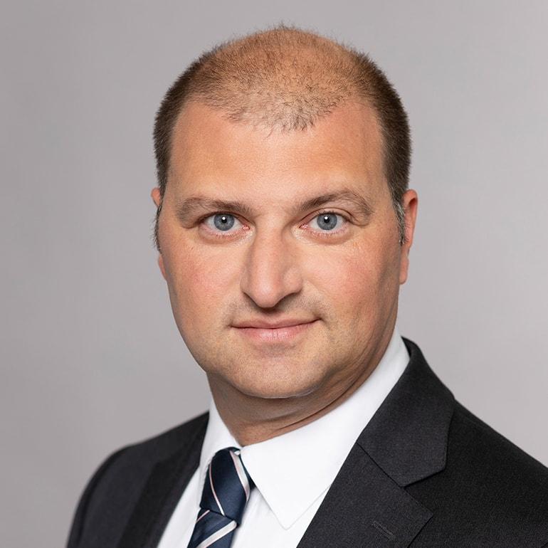 Porträt eines Mannes mit kurzem, leicht speckigem Haar und blauen Augen. Er trägt einen dunklen Anzug mit einem weißen Hemd und einer gestreiften Krawatte. Der Hintergrund ist neutral grau. Der Mann schaut direkt in die Kamera und hat einen neutralen Gesichtsausdruck.