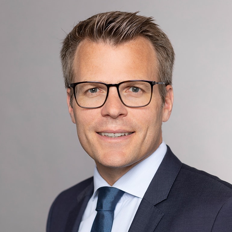 Ein lächelnder Mann mit kurzen, grauen Haaren trägt eine Brille und einen Anzug mit Krawatte. Der Hintergrund ist in sanften Grautönen gehalten. Die Bildkomposition vermittelt Professionalität und Freundlichkeit.