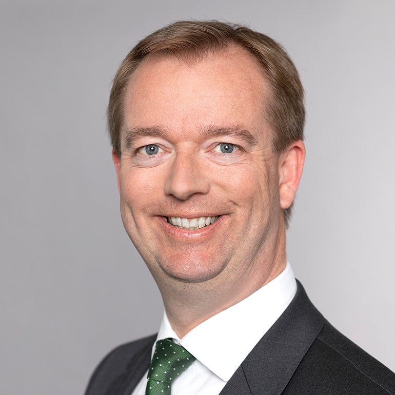 Ein lächelnder Mann mit kurzen, blond-bräunlichen Haaren trägt einen Anzug und eine Krawatte mit grünen Punkten. Er hat blaue Augen und sieht freundlich aus. Der Hintergrund ist hellgrau und unauffällig.