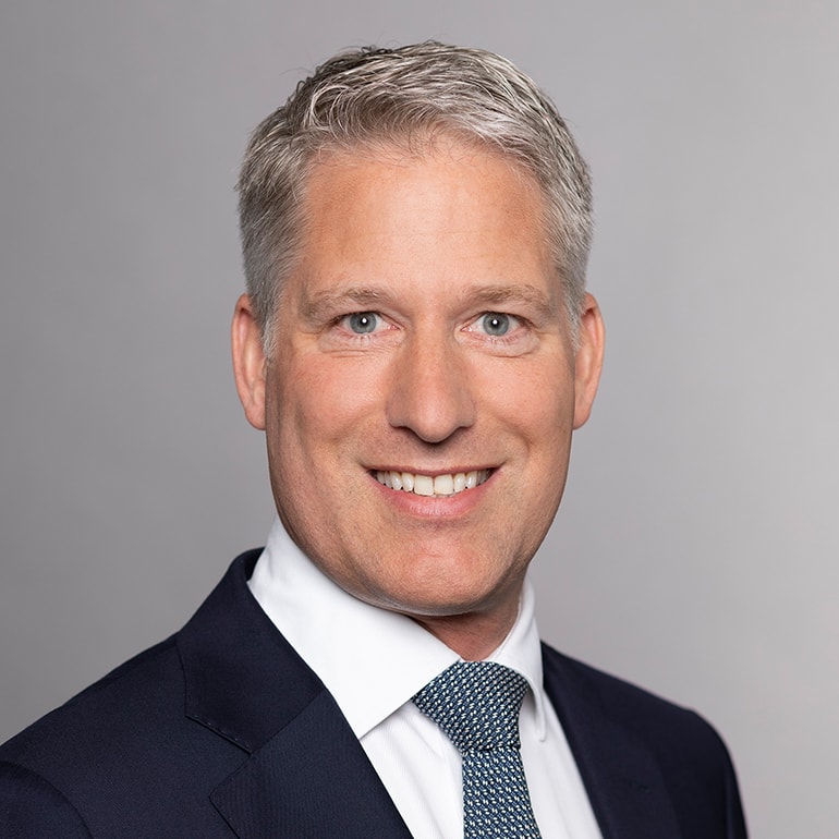 Ein professionell gekleideter Mann mit kurzen, blonden Haaren und einem freundlichen Lächeln blickt in die Kamera. Er trägt einen dunklen Anzug mit einem weißen Hemd und einer gestreiften Krawatte. Der Hintergrund ist neutral und einfarbig.