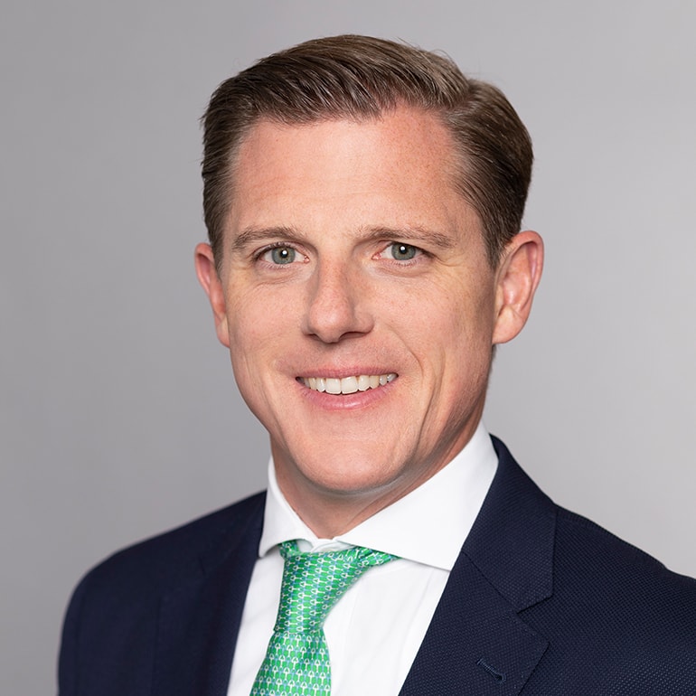 Porträt eines Mannes mit kurzem, braunem Haar und einem freundlichen Lächeln. Er trägt einen dunklen Anzug und ein grünes Oberhemd mit Krawatte. Der Hintergrund ist einfarbig grau, was den Fokus auf das Gesicht des Mannes lenkt.