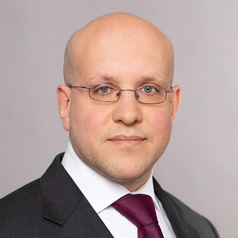 Porträt eines Mannes mit Glatze, der eine Brille trägt. Er ist im Anzug mit einer Krawatte gekleidet und schaut direkt in die Kamera. Der Hintergrund ist neutral und leicht verschwommen, um den Fokus auf das Gesicht zu legen.