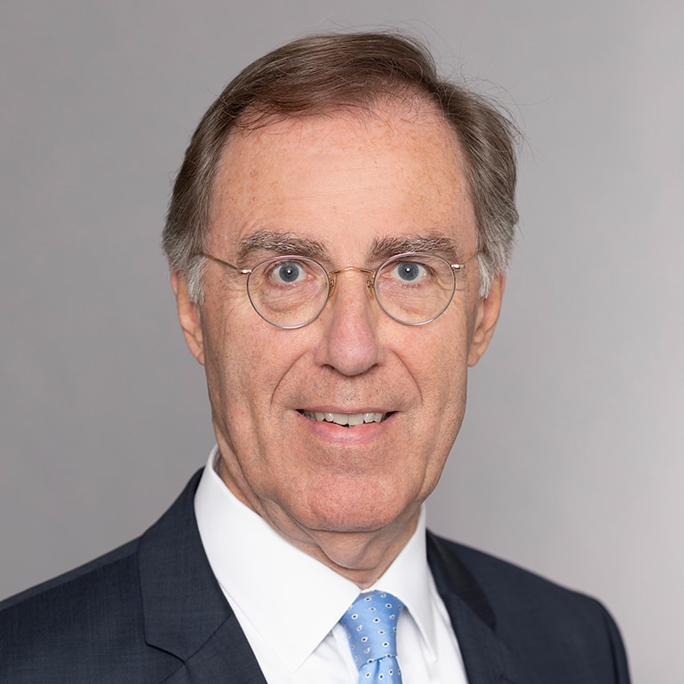 Ein älterer Mann mit Brille lächelt freundlich in die Kamera. Er trägt ein dunkelblaues Sakko und ein weißes Hemd mit einer blauen Krawatte. Der Hintergrund ist neutral und hellgrau.