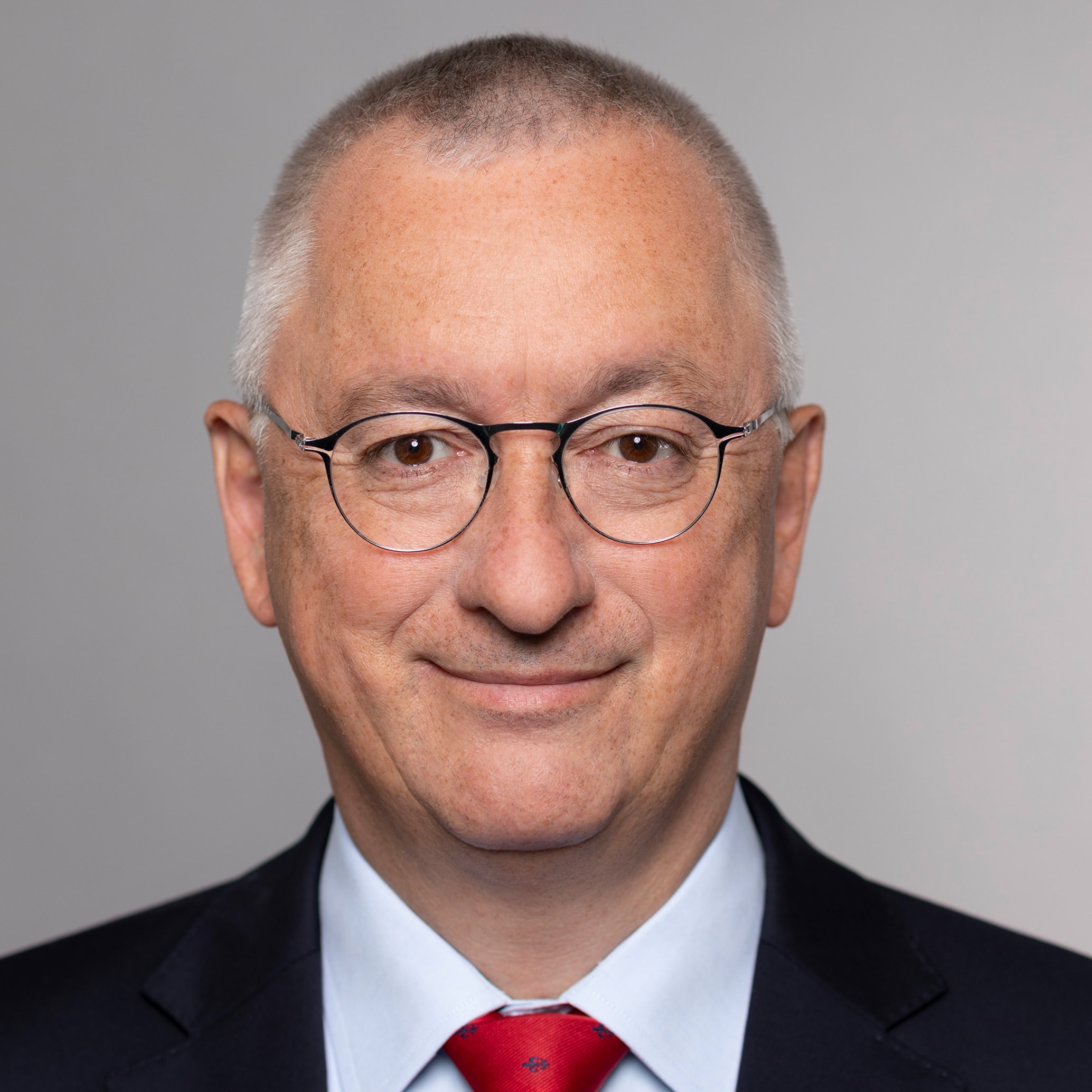 Porträt eines Mannes mit kurzem, grauem Haar, rundem Gesicht und Brille. Er trägt einen dunkelblauen Anzug mit einer hellblauen Hemd und einer roten Krawatte. Der Hintergrund ist grau. Der Mann lächelt leicht.