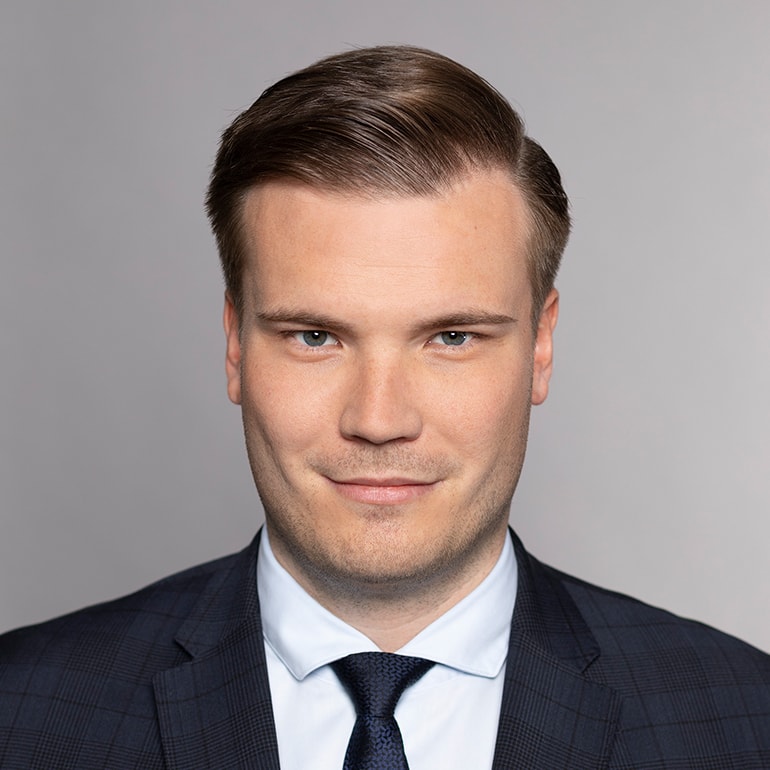 Ein junger Mann mit kurzen, gestylten Haaren und einem leichten Lächeln trägt einen dunkelblauen Anzug mit Krawatte. Er schaut direkt in die Kamera vor einem neutralen grauen Hintergrund. Seine Gesichtszüge sind freundlich und selbstbewusst.