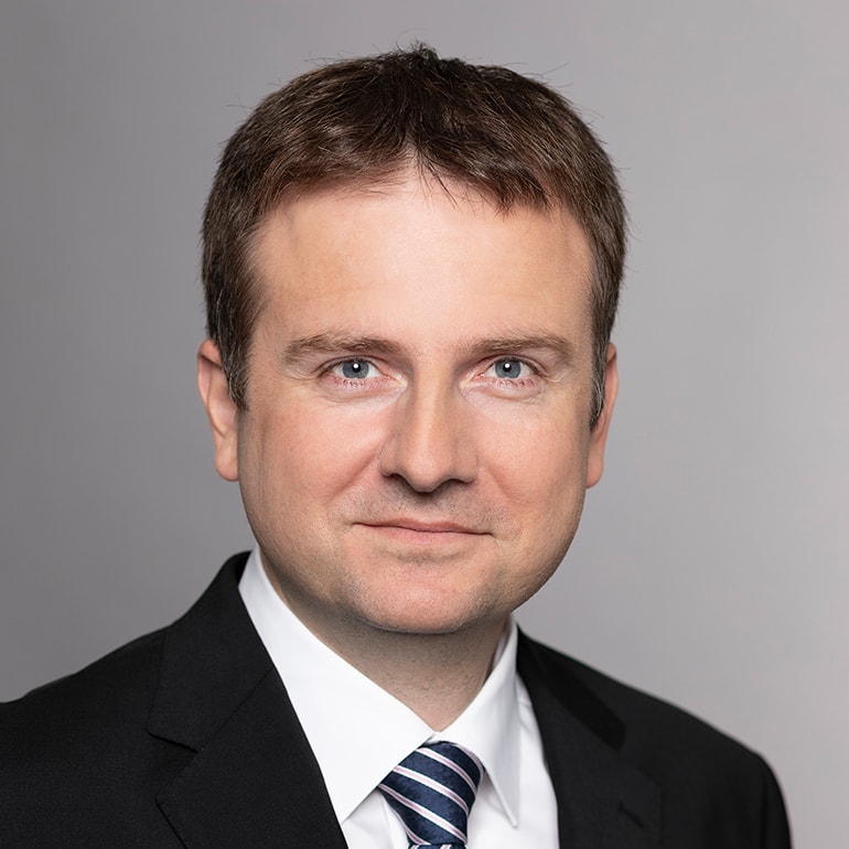 Ein seriöser Mann mit kurzer, brauner Haarfrisur trägt einen dunklen Anzug und ein gestreiftes Hemd. Er schaut direkt in die Kamera, hat einen neutralen Gesichtsausdruck und einen hellen Hintergrund.