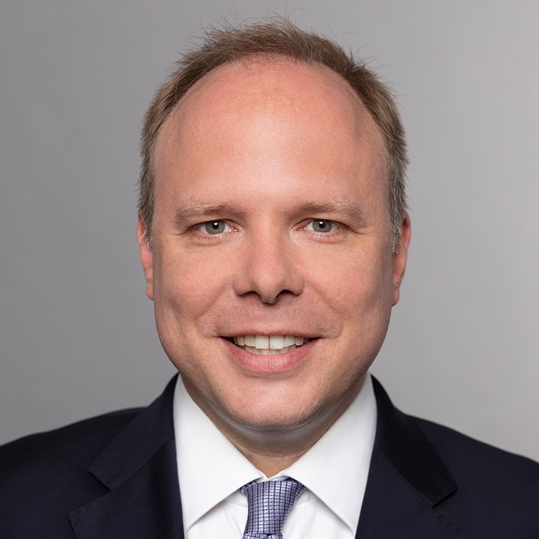 Porträt eines lächelnden Mannes mit kurzen, hellen Haaren. Er trägt einen dunkelblauen Anzug und ein weißes Hemd mit einer gestreiften Krawatte. Der Hintergrund ist hellgrau. Der Mann wirkt freundlich und professionell.