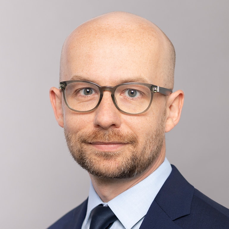 Ein Mann mit glattem Kopf und Brille lächelt in die Kamera. Er trägt einen blauen Anzug und ein hellblaues Hemd mit dunkler Krawatte. Der Hintergrund ist einfarbig und neutral.