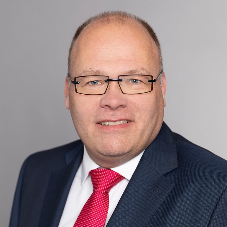 Porträt eines Mannes mit Brille, der in einem dunklen Anzug und einer roten Krawatte gekleidet ist. Er lächelt freundlich in die Kamera, vor einem hellgrauen Hintergrund.