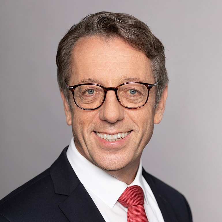 Porträt eines Mannes in einem schwarzen Anzug mit einem weißen Hemd und einer roten Krawatte. Er trägt eine Brille und lächelt leicht. Der Hintergrund ist grau. Der Mann hat kurze, graue Haare und einen freundlichen Gesichtsausdruck.