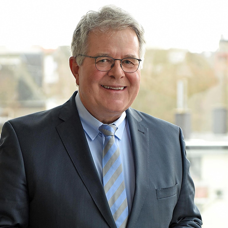 Ein lächelnder Mann mittleren Alters trägt einen dunklen Anzug mit einem hellblauen Hemd und einer gestreiften Krawatte. Er steht vor einem Fenster, durch das unscharf die Konturen einer Stadt zu sehen sind.