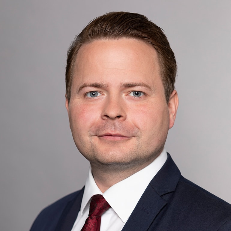 Porträt eines Mannes mit kurzen, braunen Haaren und blauen Augen. Er trägt einen dunkelblauen Anzug mit einem weißen Hemd und einer roten Krawatte. Der Hintergrund ist grau. Der Mann schaut direkt in die Kamera und hat einen neutralen Gesichtsausdruck.