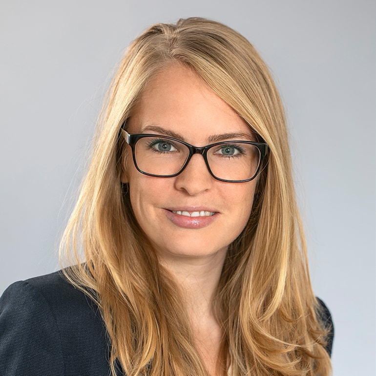 Porträt einer blonden Frau mit langen Haaren, die eine Brille trägt. Sie lächelt freundlich und trägt einen dunklen Blazer. Der Hintergrund ist hell und neutral.