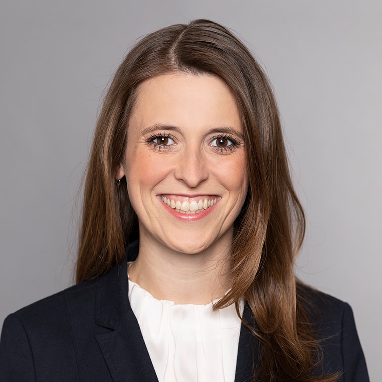 Portrait einer lächelnden Frau mit langen, braunen Haaren. Sie trägt einen dunkelblauen Blazer und ein helles Oberteil. Der Hintergrund ist einfarbig grau. Das Bild vermittelt Professionalität und Freundlichkeit.