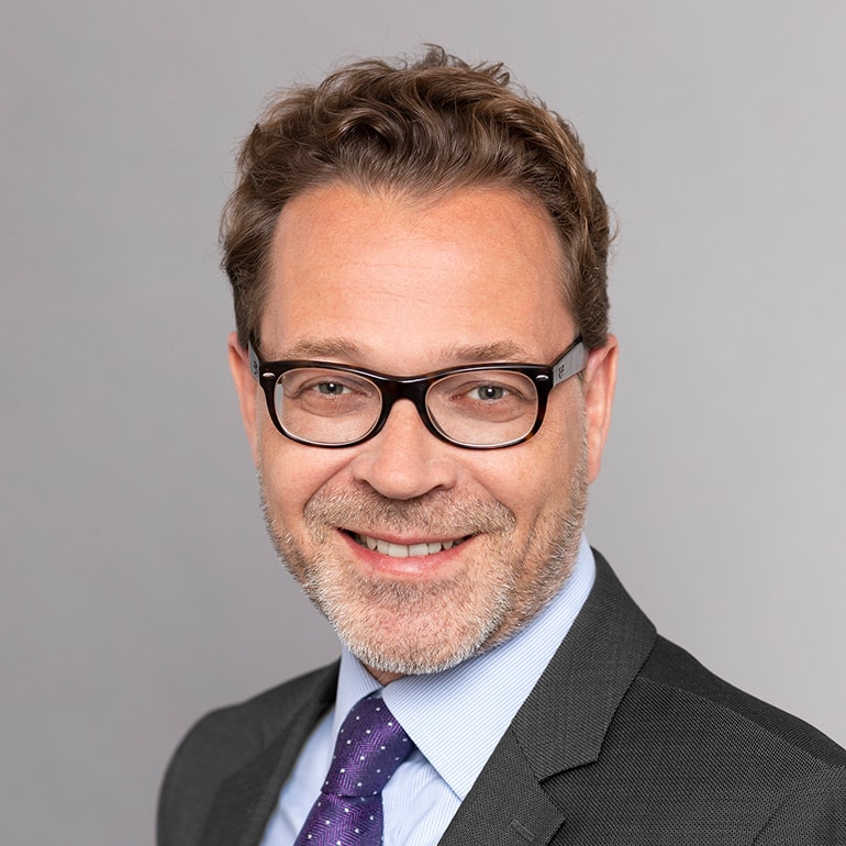 Ein lächelnder Mann mit Brille steht vor einem grauen Hintergrund. Er trägt einen dunklen Anzug und ein hellblaues Hemd mit einer lila Krawatte. Der Mann wirkt freundlich und professionell.