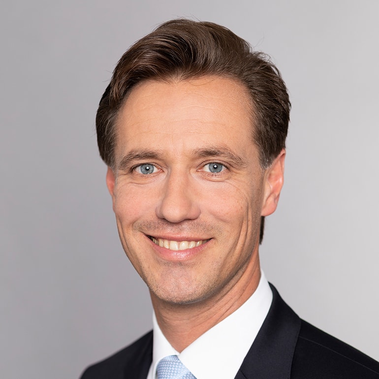 Porträt eines Mannes mit kurzen, braunen Haaren und blauem Anzug. Er hat ein freundliches Lächeln, trägt eine hellblaue Krawatte und ist vor einem grauen Hintergrund abgebildet.