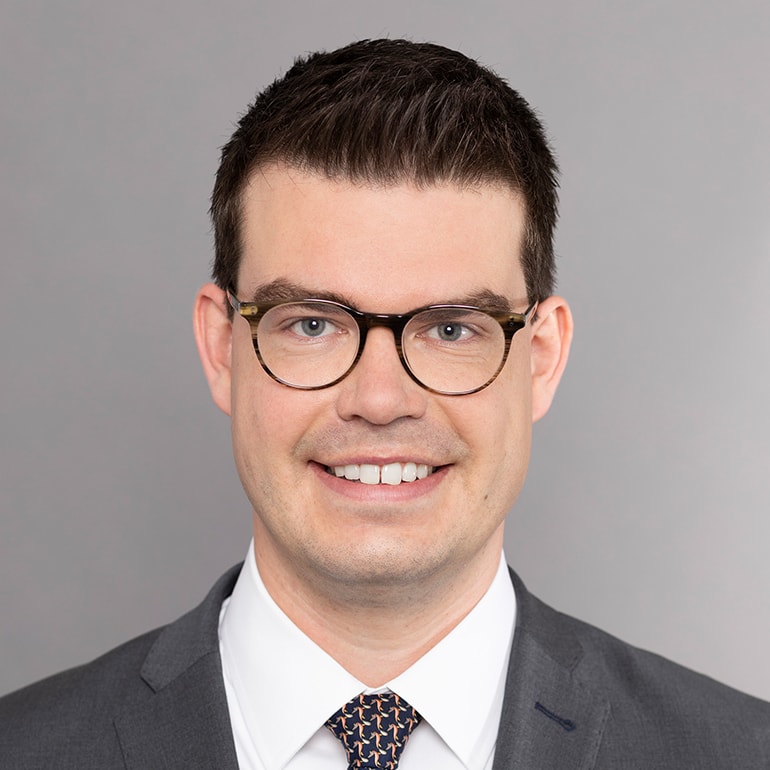 Portrait eines Mannes mit kurzen, dunklen Haaren und Brille. Er trägt einen grauen Anzug mit einem weißen Hemd und einer gemusterten Krawatte. Der Mann lächelt freundlich vor einem neutralen, hellgrauen Hintergrund.