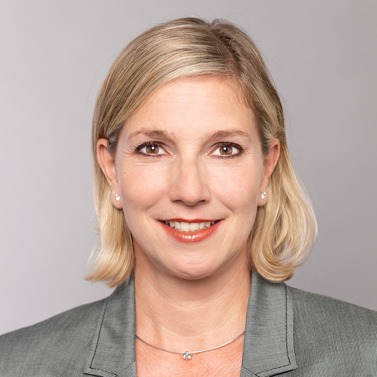 Porträt einer Frau mit blonden Haaren, die aufrecht in einem grauen Blazer posiert. Sie hat ein freundliches Lächeln und trägt eine dezente Kette. Der Hintergrund ist neutral und hell, wodurch der Fokus auf ihrem Gesicht liegt.