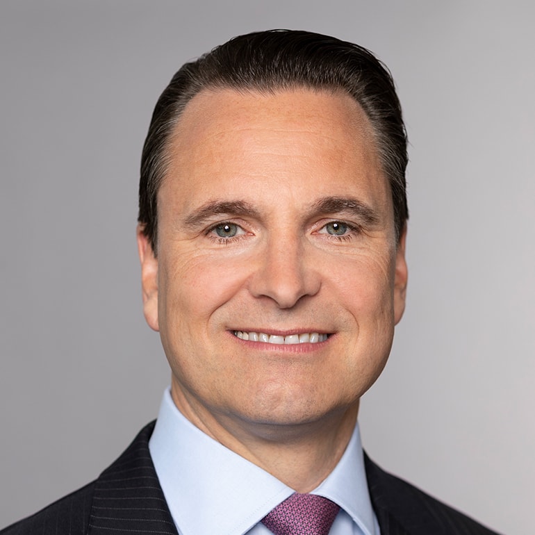 Ein lächelnder Mann mit kurzen, braunen Haaren trägt einen dunklen Anzug und ein hellblaues Hemd mit einer lila Krawatte. Der Hintergrund ist grau. Sein Gesicht drückt Selbstbewusstsein und Freundlichkeit aus.