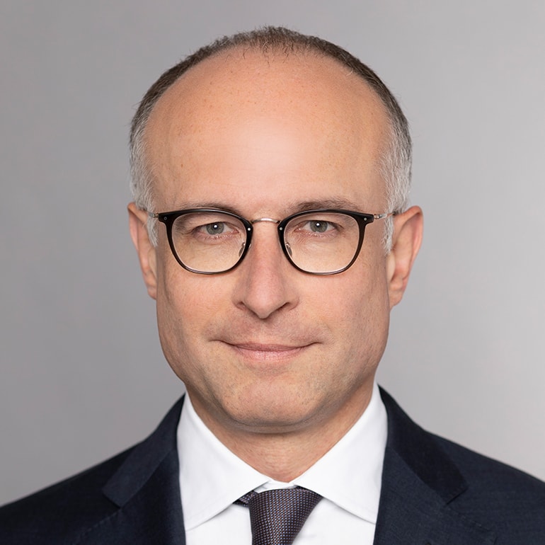 Porträt eines Mannes mit Brille, der einen Anzug und eine Krawatte trägt. Er hat kurzes, grau meliertes Haar und blickt direkt in die Kamera. Der Hintergrund ist neutral und unauffällig, um den Fokus auf das Gesicht zu lenken.