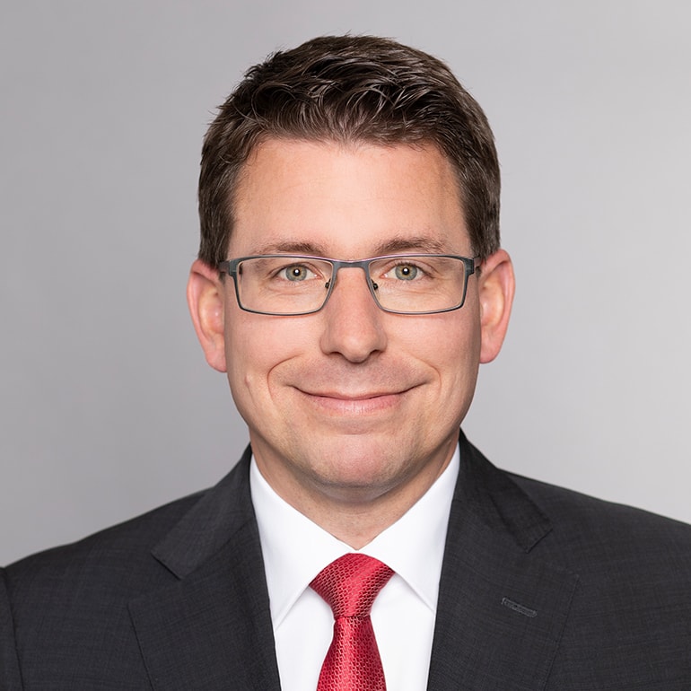 Porträt eines lächelnden Mannes mit Brille, Mittellange braune Haare, im Anzug und einer roten Krawatte. Der Hintergrund ist neutral und grau.