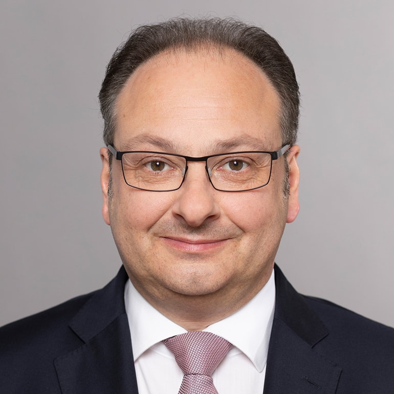 Porträt eines lächelnden Mannes mit Brille. Er trägt einen dunklen Anzug und ein helles Hemd mit einer gemusterten Krawatte. Der Hintergrund ist einfarbig grau. Das Gesicht ist freundlich und vermittelt Professionalität.