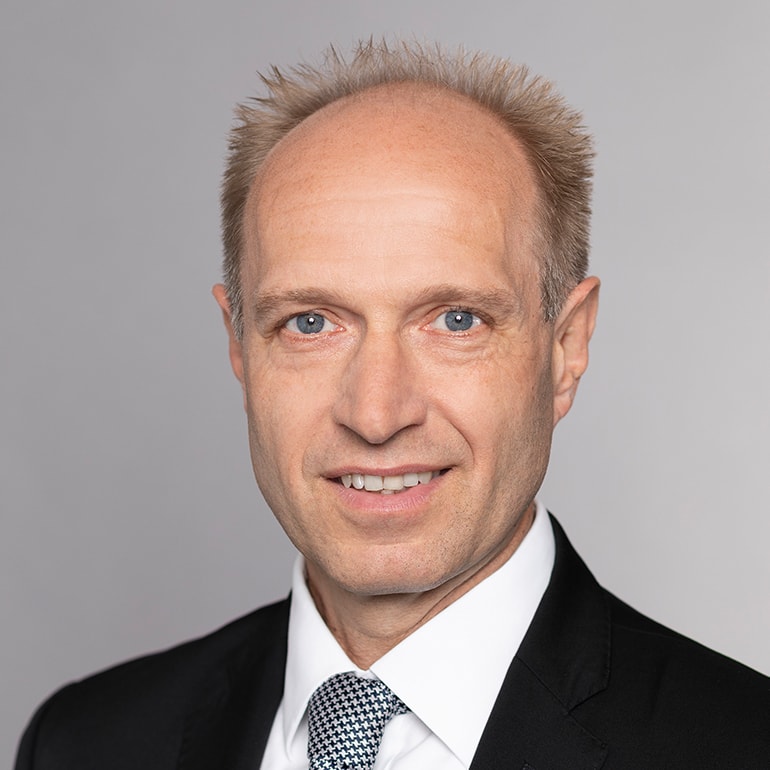 Porträt eines Mannes mit blonden, kurzen Haaren und einem freundlichen Lächeln. Er trägt einen schwarzen Anzug mit einem karrierten Krawatte und hat blaue Augen. Der Hintergrund ist grau und neutral gehalten.