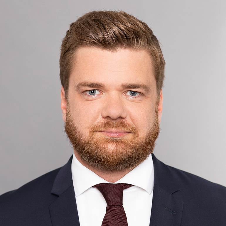 Ein männlicher Geschäftsmann mit kurzem, gestylt Haar und einem voluminösen Bart blickt direkt in die Kamera. Er trägt einen dunkelblauen Anzug und eine weiße Hemdbluse mit einer rotbraunen Krawatte, vor einem neutralen grauen Hintergrund.