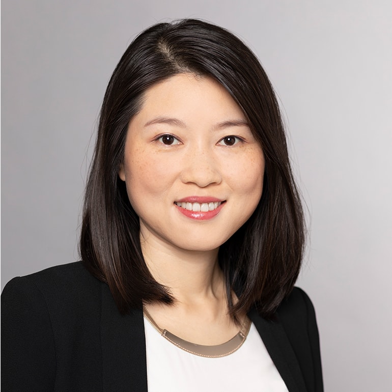 Porträt einer Frau mit schulterlangem, glattem Haar. Sie trägt ein schwarzes Blazer-Jacket und eine helle Bluse. Die Frau lächelt freundlich in die Kamera, der Hintergrund ist hellgrau.