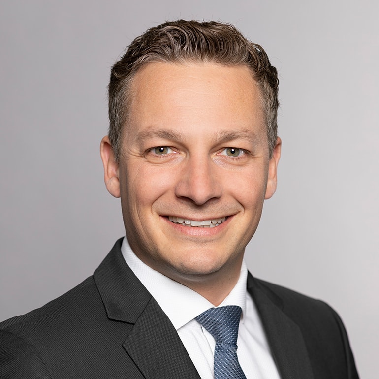 Das Bild zeigt einen lächelnden Mann mit kurzen, braunen Haaren. Er trägt einen schwarzen Anzug und eine hellblaue Krawatte. Der Hintergrund ist grau. Die Darstellung vermittelt einen professionellen und freundlichen Eindruck.
