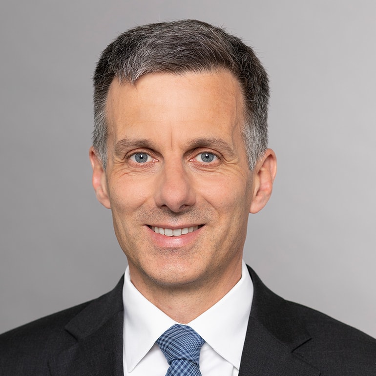 Ein mittelalter Mann mit kurzen, grauen Haaren lächelt in die Kamera. Er trägt einen dunkelgrauen Anzug und eine helle Hemd mit einer blauen Krawatte. Der Hintergrund ist hellgrau.