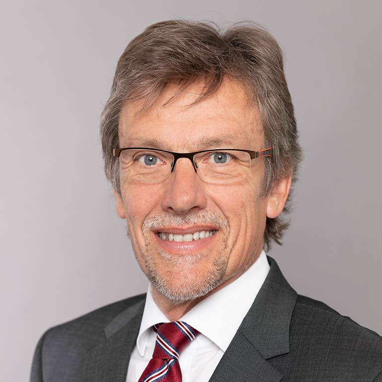 Portrait eines lächelnden Mannes mit Brille, mittellangen, grauen Haaren. Er trägt einen dunklen Anzug und eine gestreifte Krawatte. Der Hintergrund ist grau und neutral gehalten.