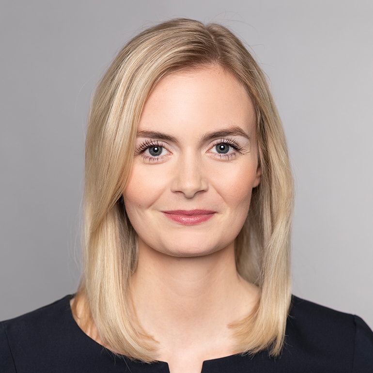 Ein Porträt einer Frau mit blonden, schulterlangen Haaren, die direkt in die Kamera lächelt. Sie trägt ein dunkles Oberteil. Der Hintergrund ist grau, was ihr Gesicht betont. Ihre Augen sind hell und freundlich.