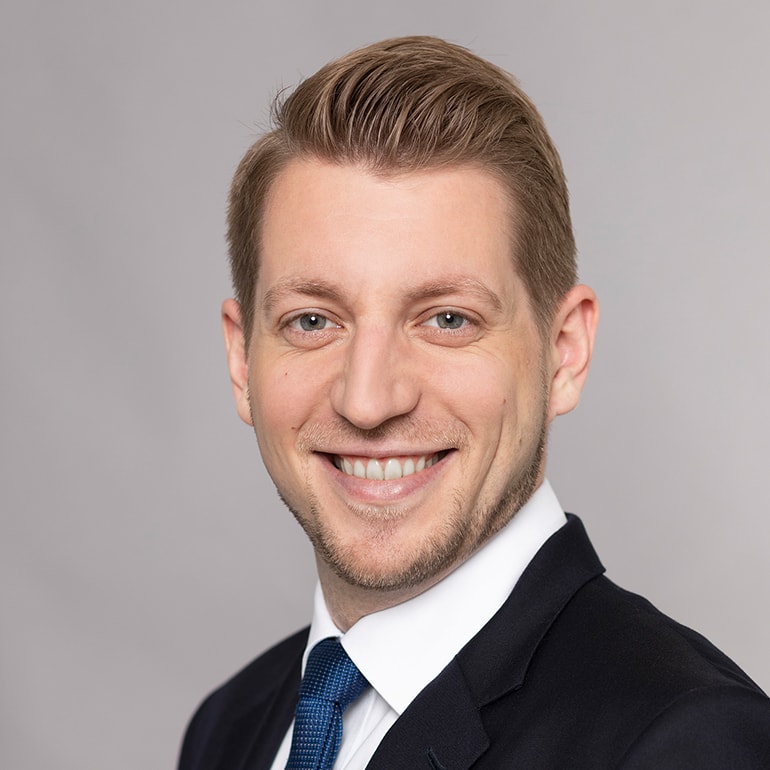 Ein junger Mann mit blonden Haaren und einem freundlichen Lächeln trägt einen schwarzen Anzug und eine blaue Krawatte. Der Hintergrund ist grau. Die Bildkomposition vermittelt Professionalität und Selbstbewusstsein.