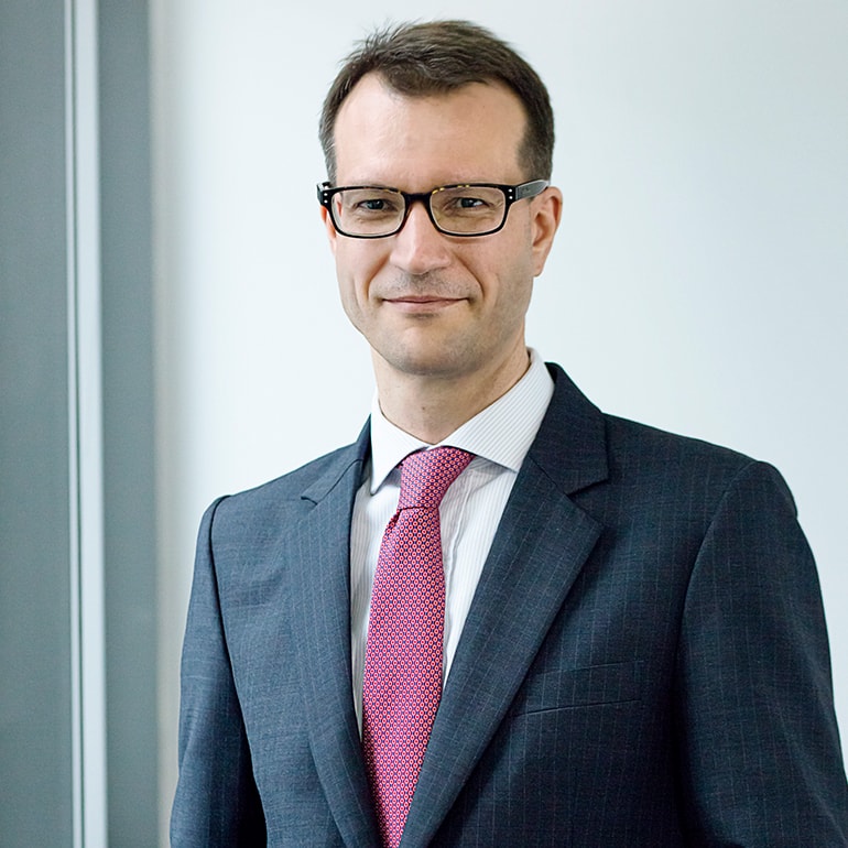 Ein Mann in einem dunkelgrauen Anzug mit kariertem Muster und einer roten Krawatte steht vor einer hellen Wand. Er trägt eine Brille und lächelt leicht, während er in die Kamera schaut.