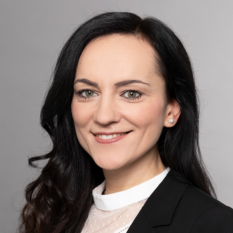 Porträt einer lächelnden Frau mit langen, schwarzen Haaren, die einen dunklen Blazer und ein helles Oberteil trägt. Der Hintergrund ist grau. Die Frau zeigt einen freundlichen Ausdruck und hat ihre Haare offen.