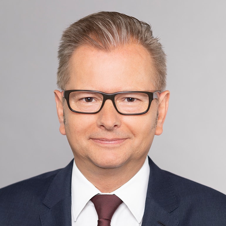 Porträt eines Mannes mit grauen Haaren und Brille, der in einem Anzug mit Krawatte vor einem neutralen, hellgrauen Hintergrund lächelt. Er wirkt freundlich und professionell.