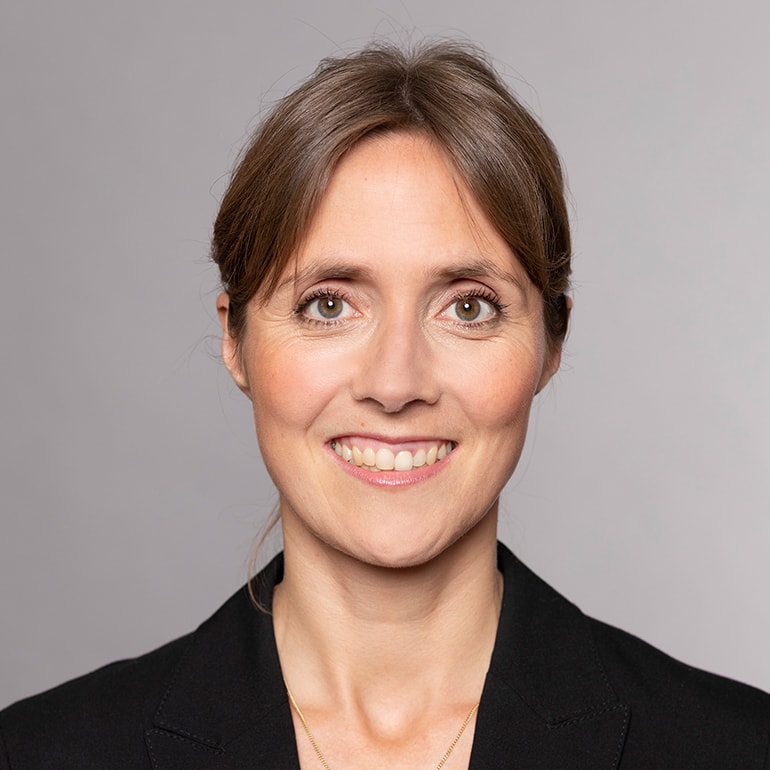 Porträt einer lächelnden Frau mit mittellangen, braunen Haaren. Sie trägt eine schwarze Jacke und hat einen freundlichen Gesichtsausdruck. Der Hintergrund ist einfarbig und neutral gehalten.