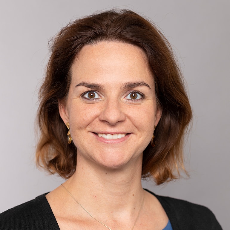 Porträt einer Frau mit braunen, schulterlangen Haaren und einem freundlichen Lächeln. Sie trägt goldene Ohrringe und eine schwarze Bluse. Der Hintergrund ist grau und unauffällig.