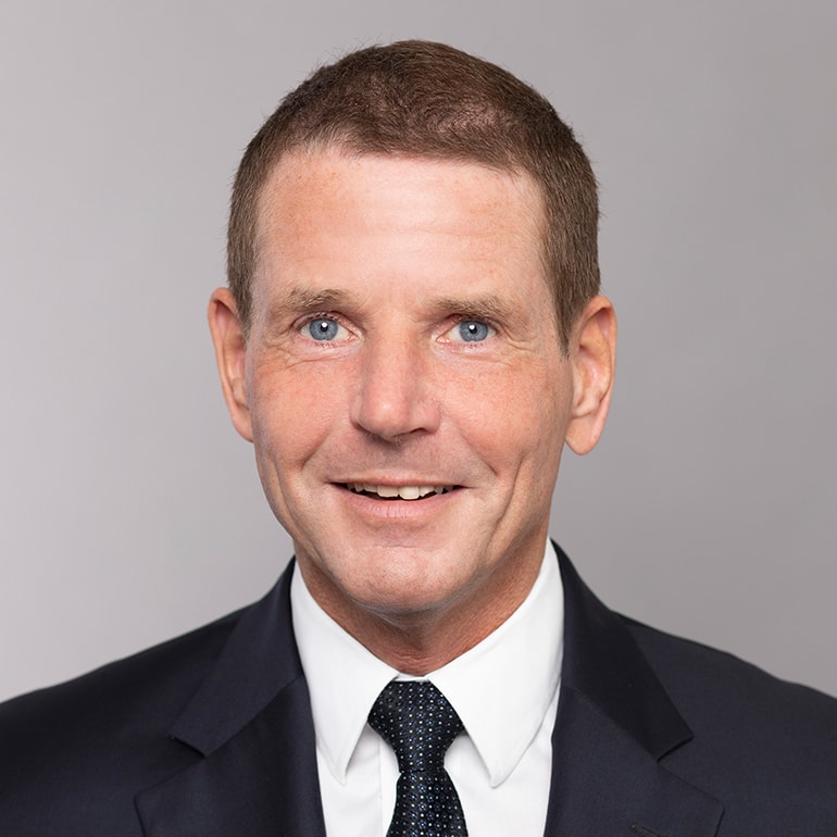 Porträt eines Mannes mit kurzem Haar und blauen Augen, der freundlich in die Kamera lächelt. Er trägt einen dunklen Anzug mit weißem Hemd und einer gemusterten Krawatte. Der Hintergrund ist hell und schlichter Farbton.