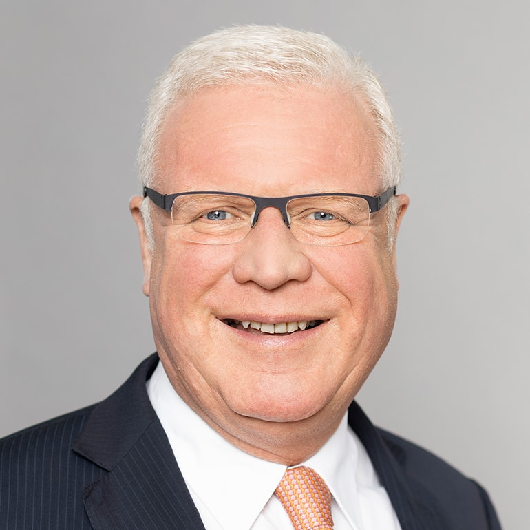 Ein lächelnder Mann mit grauem Haar trägt Brille und einen Anzug mit einem orangefarbenen Krawatte. Der Hintergrund ist grau und einfach gehalten, was den Fokus auf sein freundliches Gesicht lenkt.